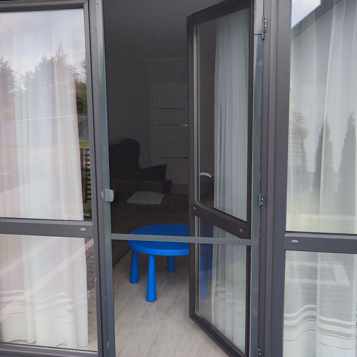 Otwarta moskitiera ramkowa w kolorze grafitowym, widok z zewnątrz, na podłodze wewnątrz widoczny niebieski stolik dziecięcy.