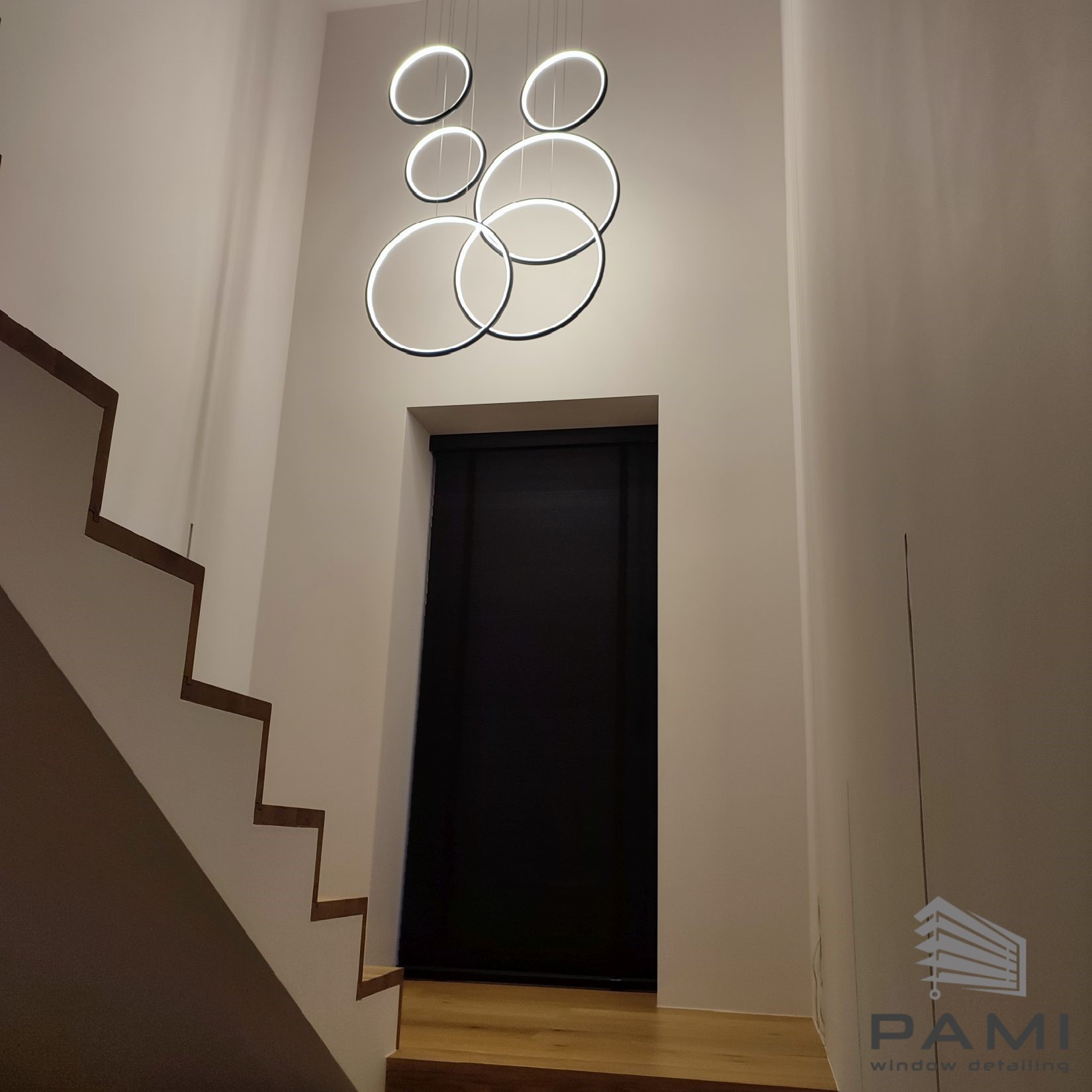 Ciemna roleta wnękowa, widoczna z dołu schodów, oświetlona nowoczesną lampą z okręgów LED, obok logo firmy 'PAMI window detailing'.