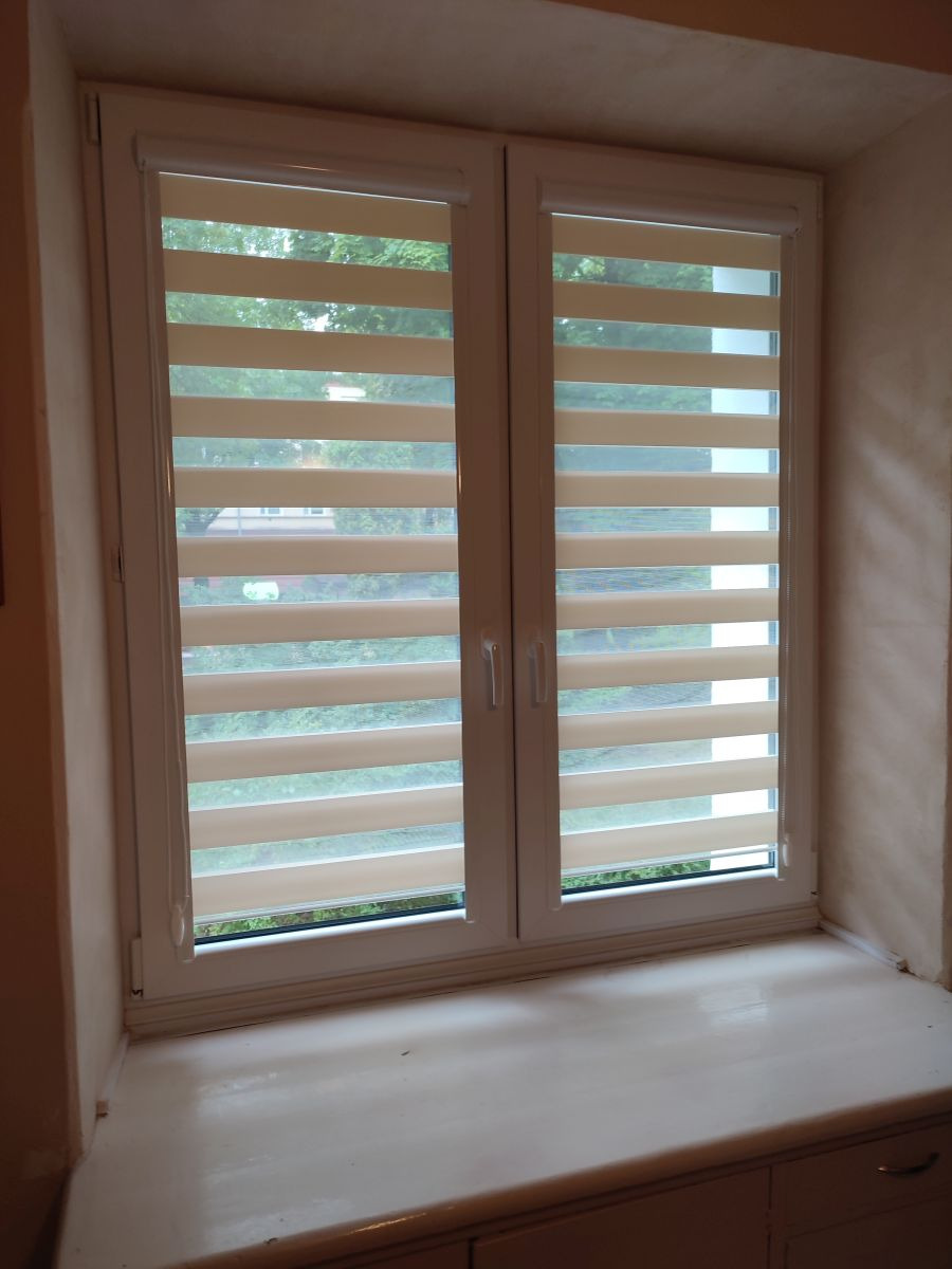 Okno z podwójnymi, poziomymi roletami w kolorze kremowym i białym, widok na zieleń za oknem, jasny parapet.