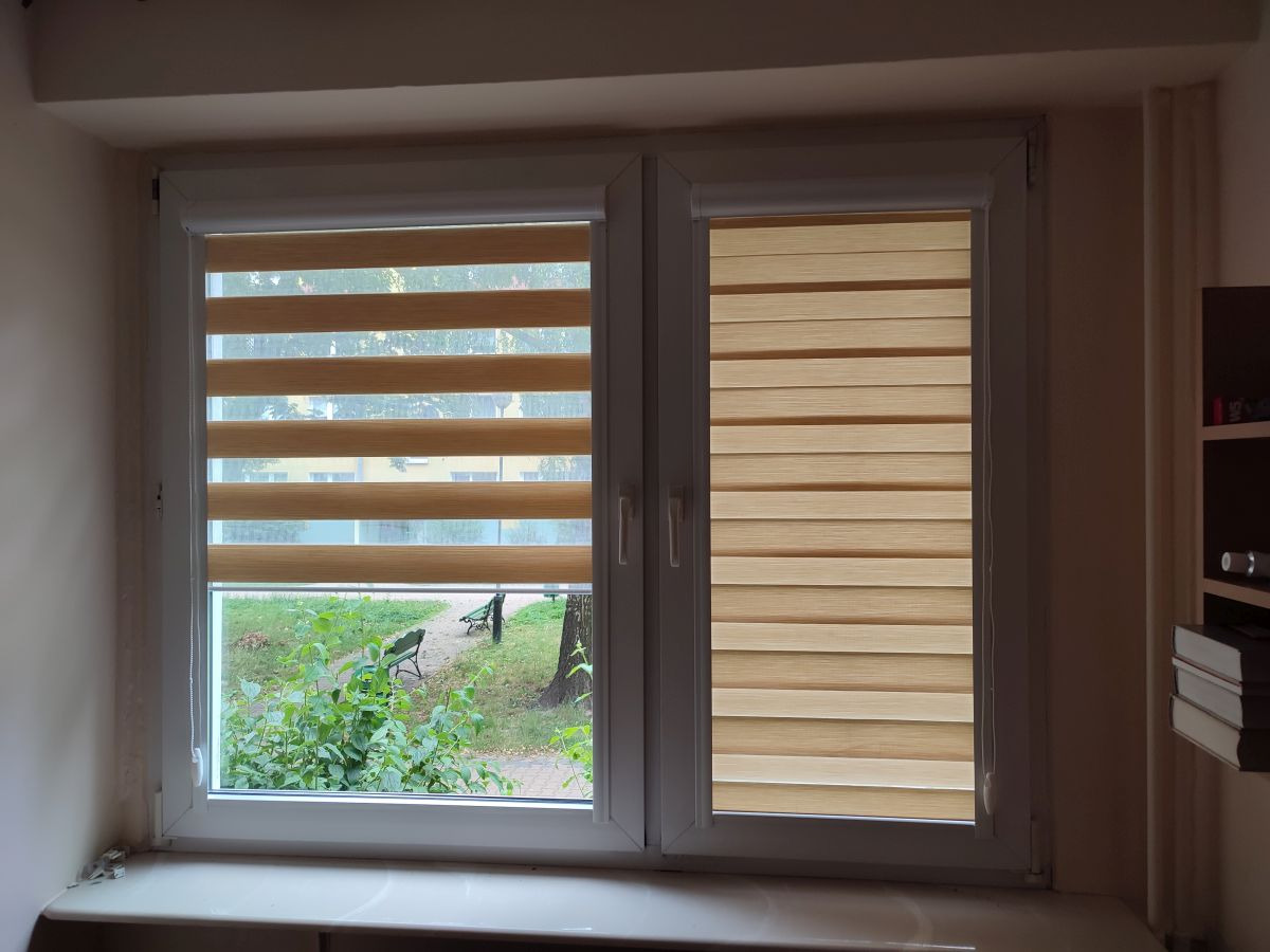 Okno z dwoma skrzydłami, na jednym z nich roleta dzień-noc z widokiem na zieleń parkową, na drugim skrzydle roleta całkowicie zasłonięta.