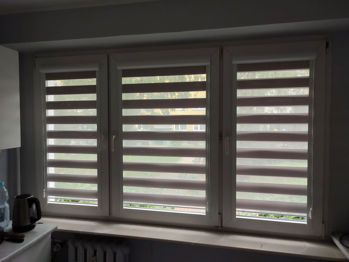 Trzy okna z zamontowanymi roletami dzień i noc z poziomymi pasami, widok z wnętrza pomieszczenia, na parapecie czajnik elektryczny i butelka wody.