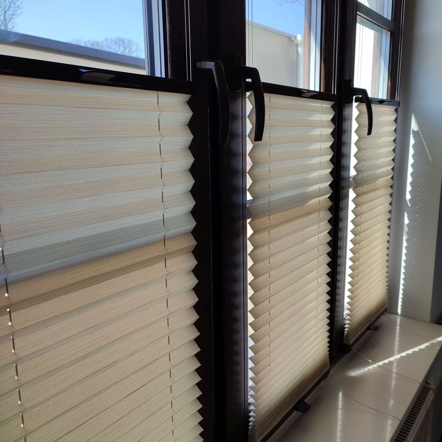 Zamknięte, kremowe, plisowane osłony okienne w brązowych ramach, z widocznymi uchwytami, zamontowane w oknie, z jasnym światłem słonecznym wpadającym do wnętrza.