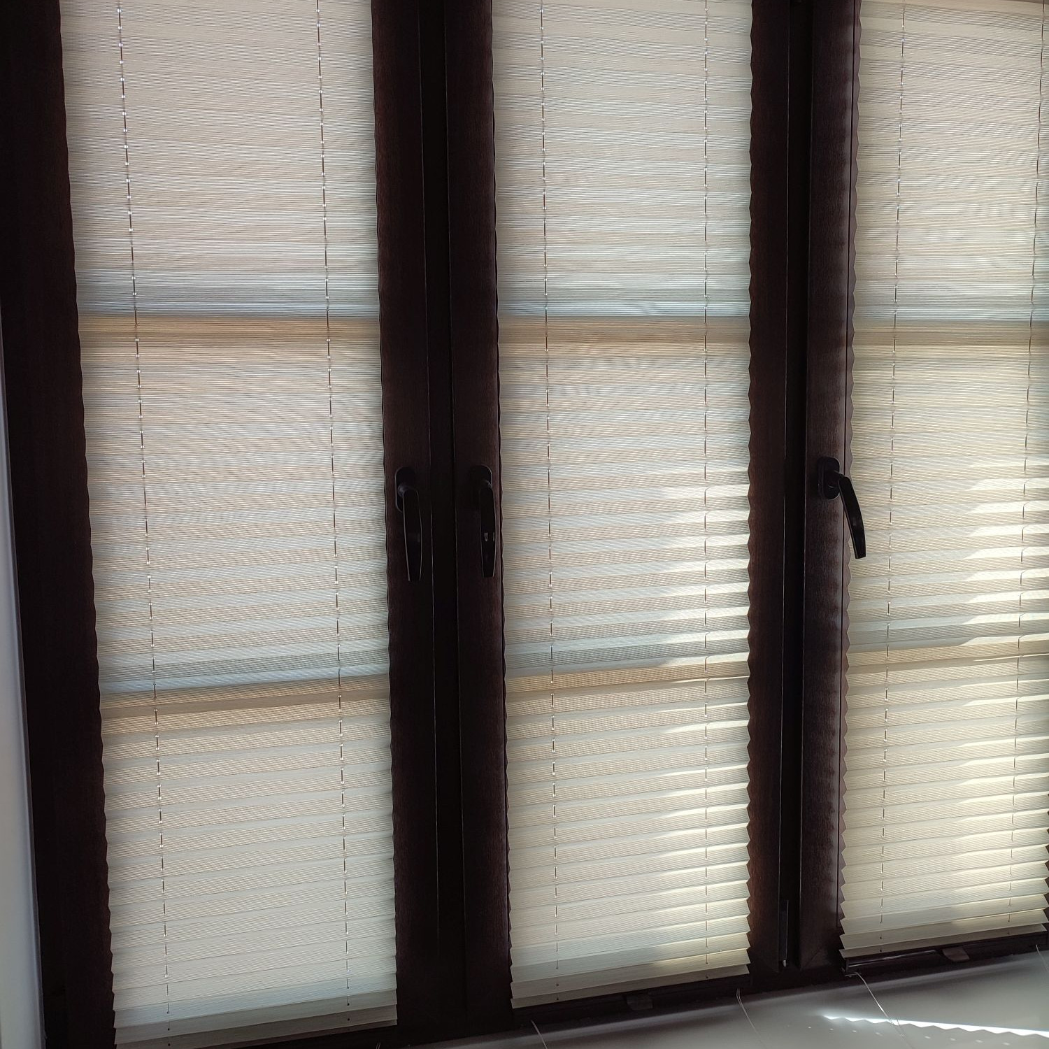 Beżowe, poziome przesłony okienne zamontowane w oknie z ciemnobrązowymi ramami, widoczne uchwyty okienne.