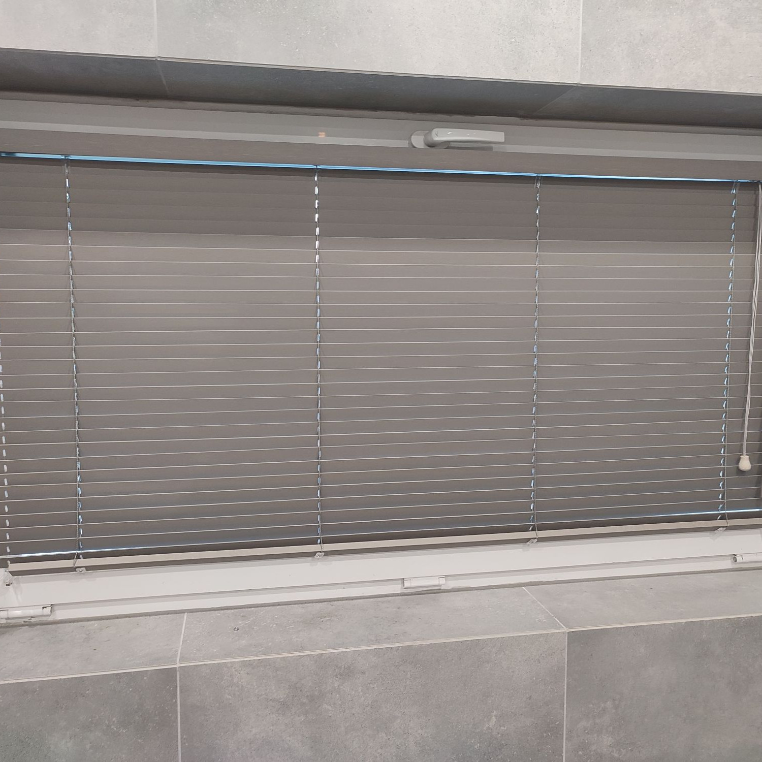 Zamknięte szare żaluzje aluminiowe w oknie, widoczne z bliska, z fragmentem parapetu wyłożonego szarymi płytkami.
