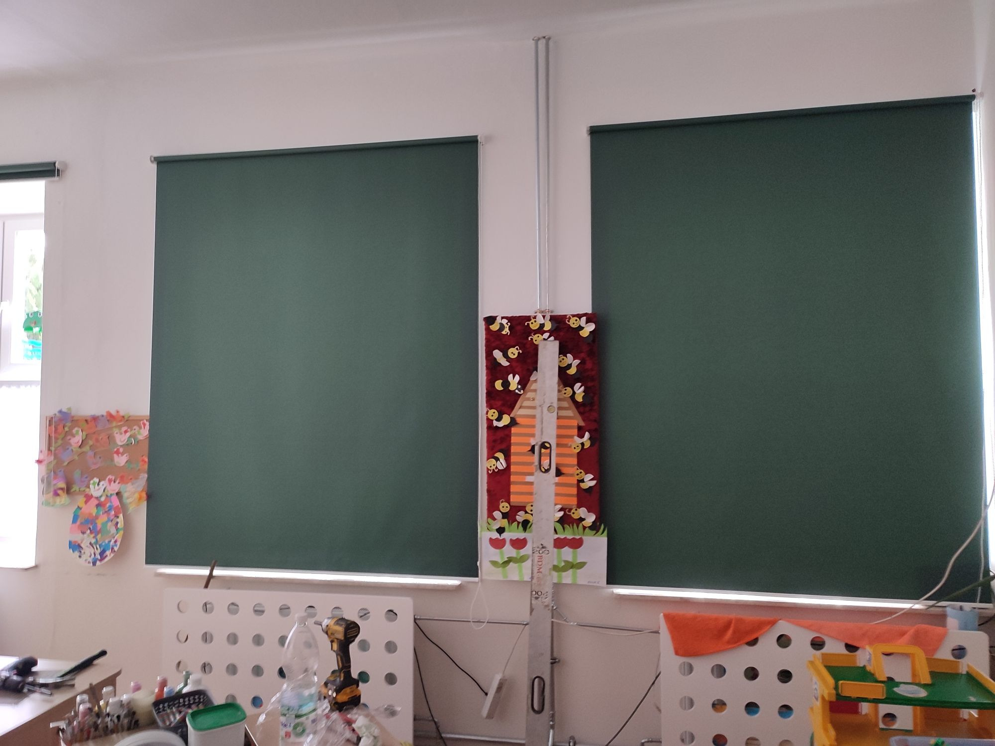 Dwa zielone zwinięte ekrany okienne w pokoju dziecięcym, z dekoracją na ścianie pomiędzy nimi i zabawkami poniżej.