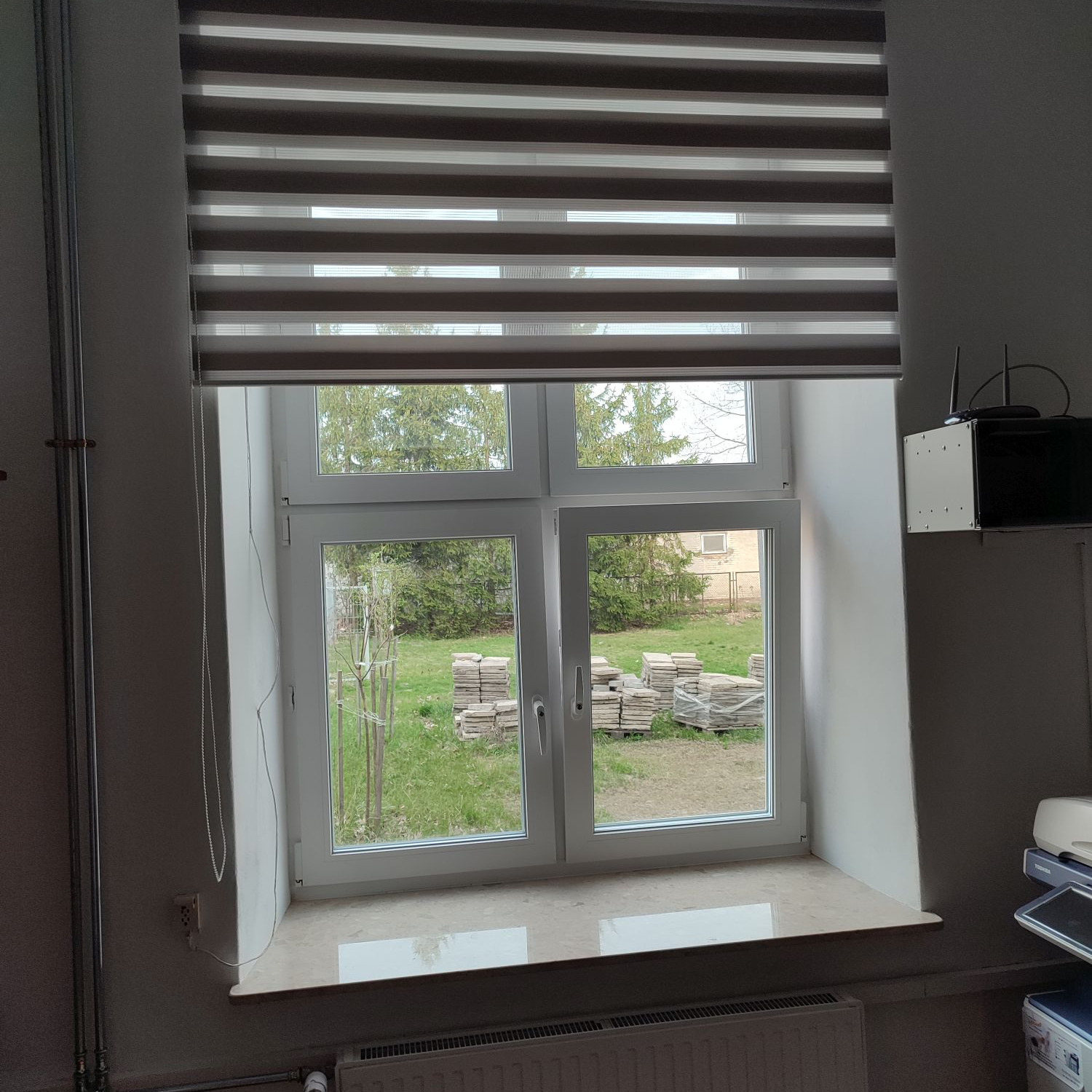 Okno z zamontowaną roletą dzień-noc w odcieniach brązu i beżu, widok na ogród z ułożonymi płytami chodnikowymi.