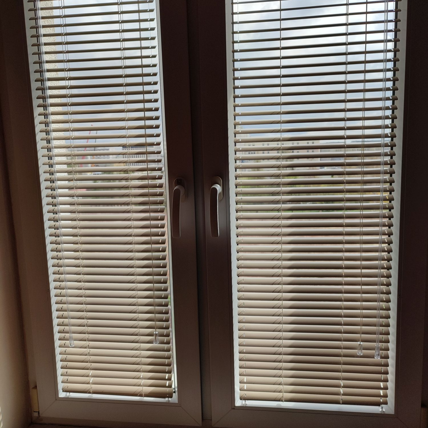 Zamknięte, poziome, beżowe listwy w aluminiowych żaluzjach okiennych, widoczne przezroczyste sznurki do regulacji