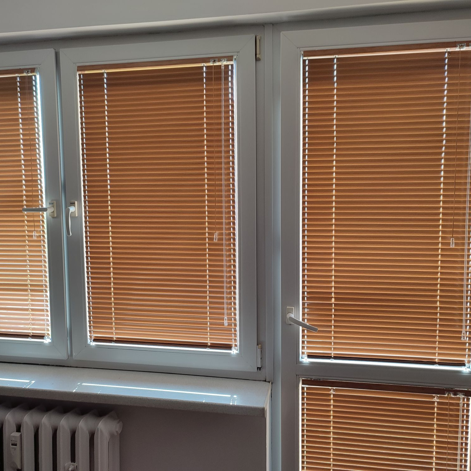 Okna z brązowymi żaluzjami aluminiowymi, widoczne fragmenty grzejnika pod parapetem, wnętrze pomieszczenia oświetlone naturalnym światłem.