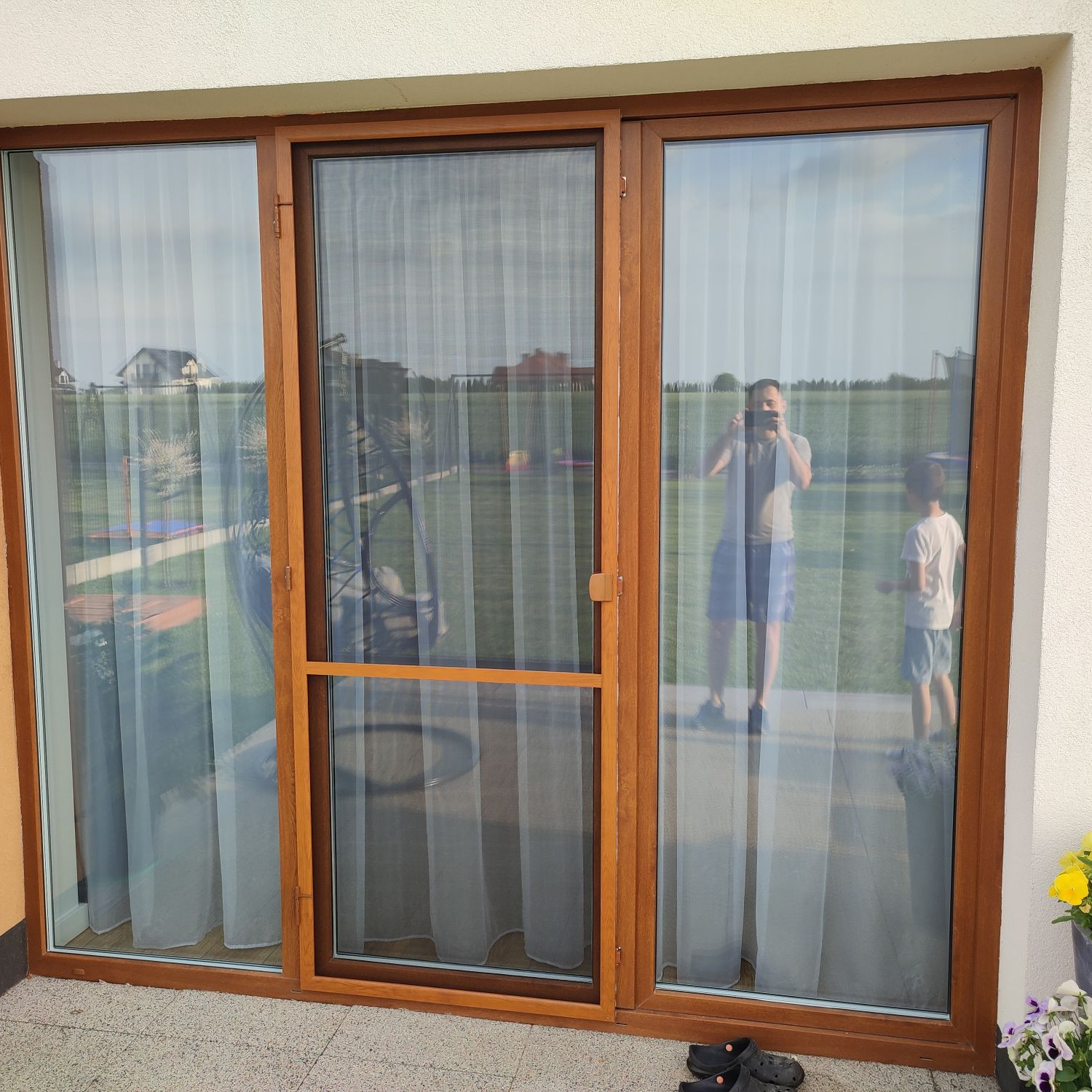 Brązowe okno tarasowe z zamontowaną siatką przeciw owadom, widoczne odbicie osoby robiącej zdjęcie i dziecka, na podłodze leżą buty.