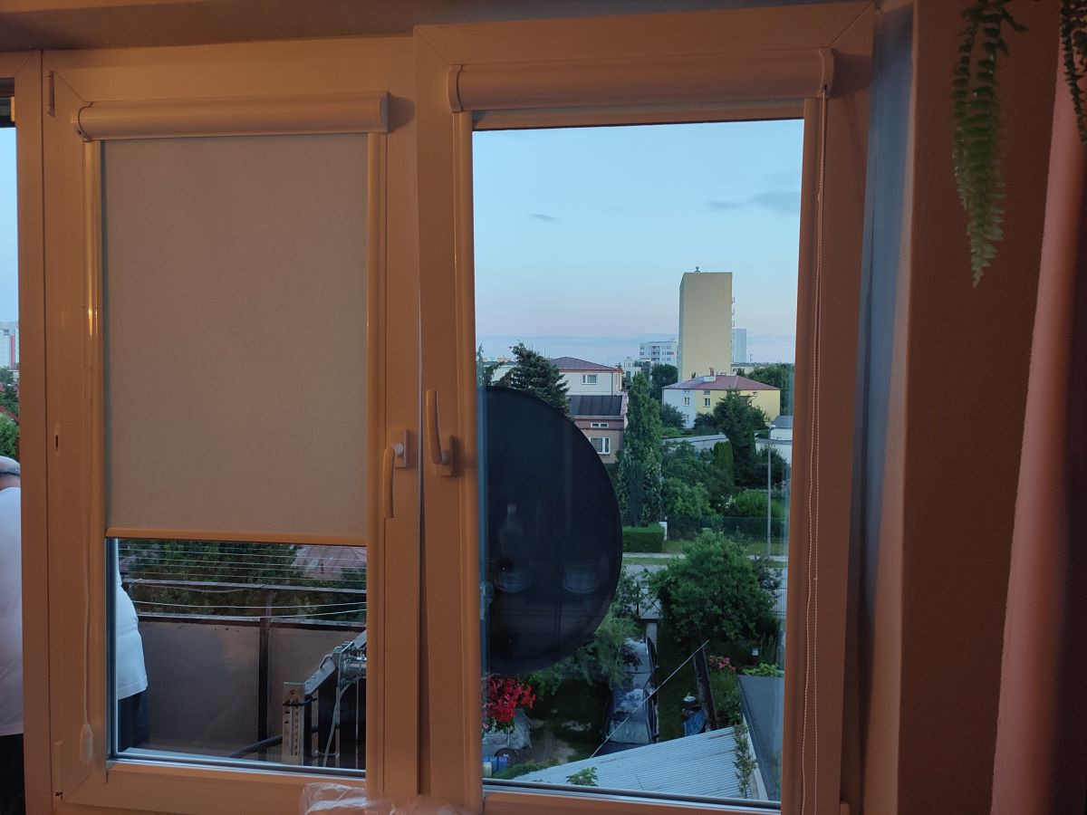 Okno z częściowo opuszczoną roletą materiałową w kolorze jasnoszarym, widok na osiedle zza okna, fragment zasłony po prawej stronie.