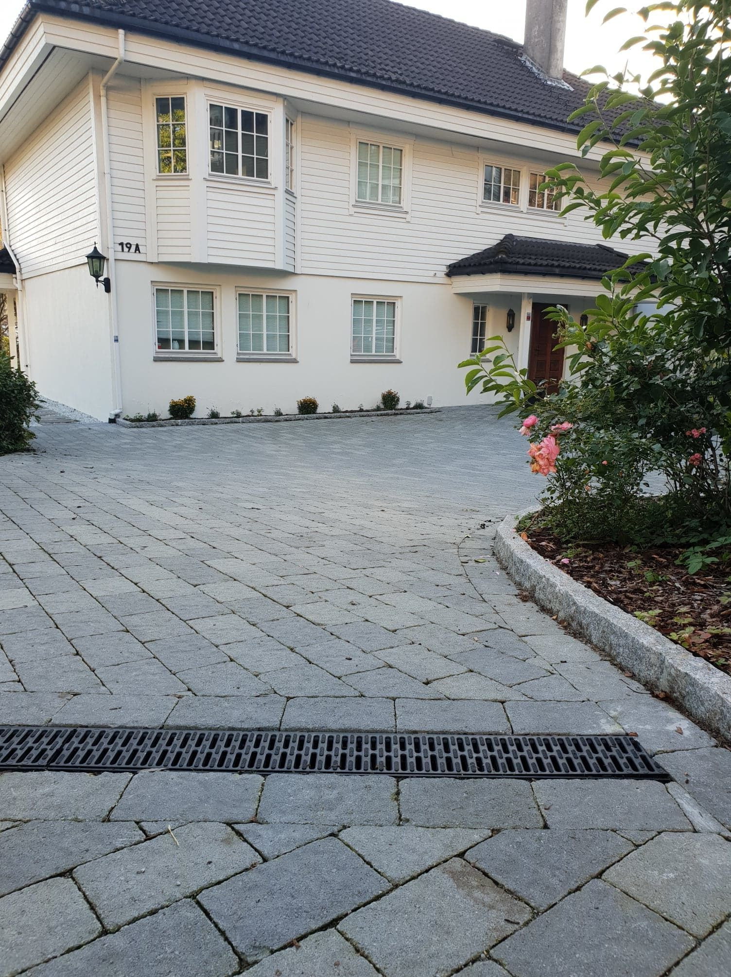 Ułożona szara kostka brukowa na podjeździe przed białym domem z numerem 19A, widoczna czarna kratka odpływowa, obrzeże z jasnego kamienia, krzew z różowymi kwiatami.