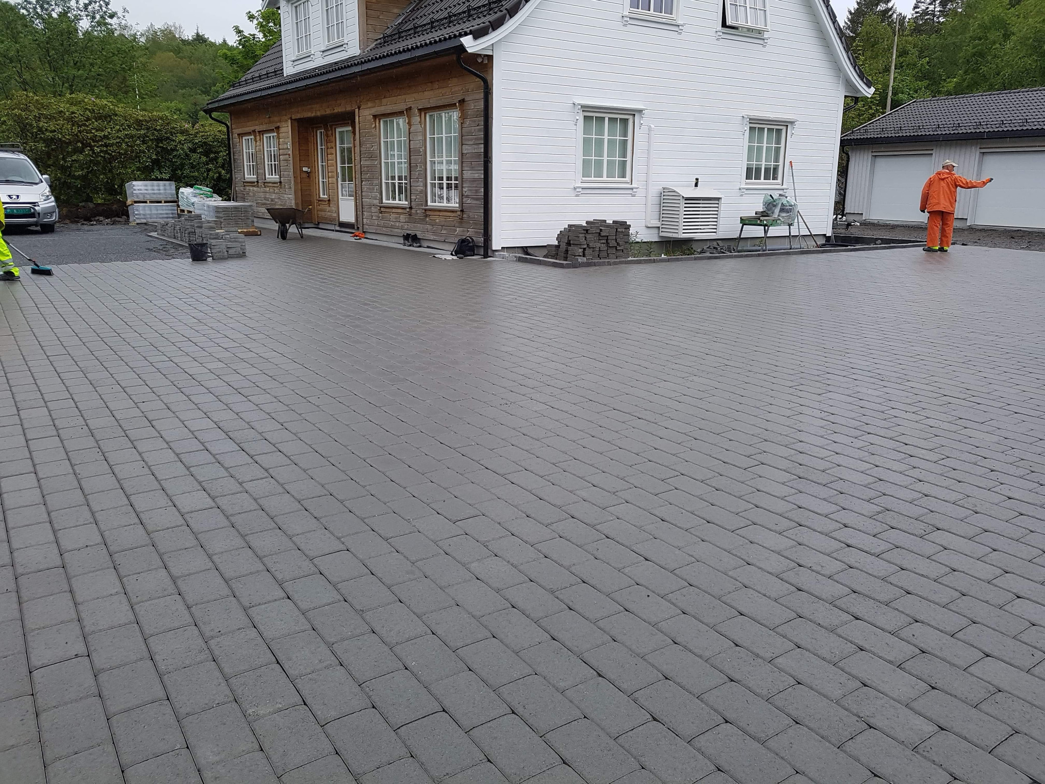 Ułożona szara kostka brukowa na podjeździe przed domem z drewnianą i białą elewacją, widoczny pracownik w pomarańczowym kombinezonie oraz zapasy kostki i narzędzia.