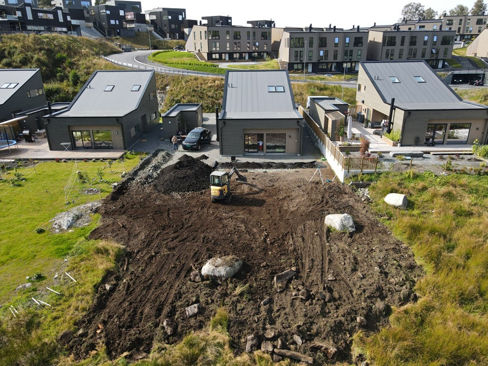 Mini koparka wyrównuje teren pod budowę kostki brukowej na posesji z nowoczesnymi domami w tle, widoczne duże kamienie i przygotowany grunt.