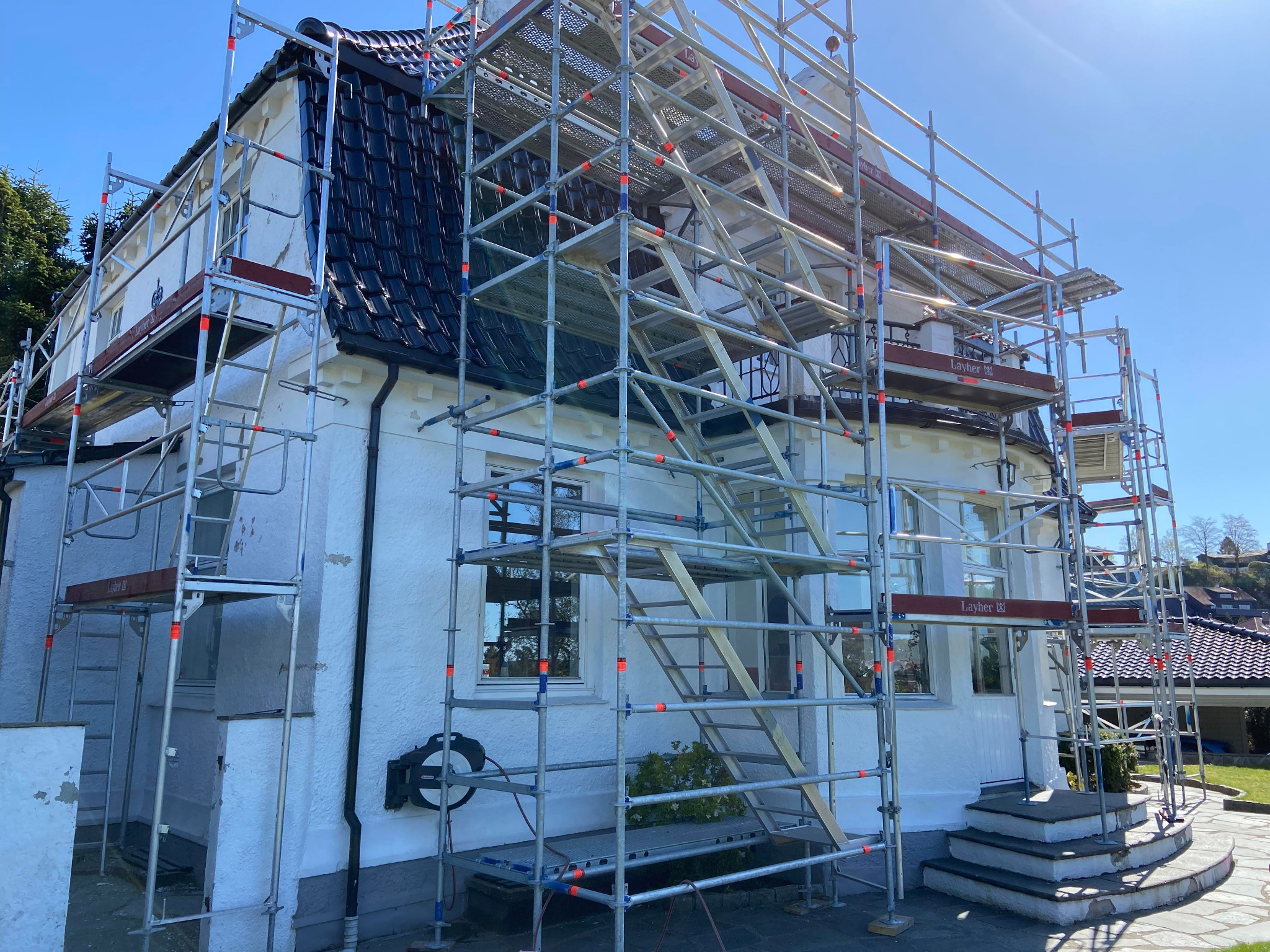 Biały dom jednorodzinny w trakcie remontu, otoczony rusztowaniami Layher z widocznymi śladami prac na elewacji i nową, błyszczącą dachówką.