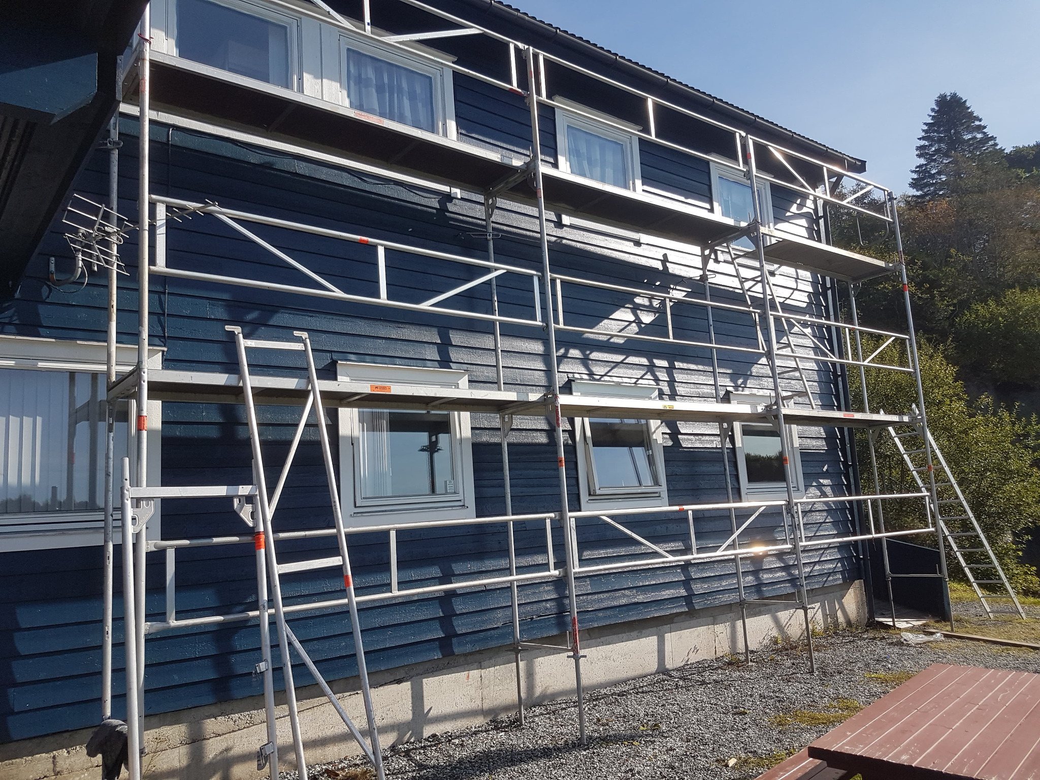 Dom z ciemnoniebieską drewnianą elewacją w trakcie malowania, rusztowanie rozstawione wzdłuż ściany, widoczne okna z białymi ramami i fragment anteny.