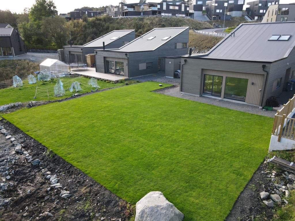 Widok z góry na zadbane trawniki przed nowoczesnymi domami z ciemnymi elewacjami i szarymi dachami. W tle widać inne budynki na wzgórzu.