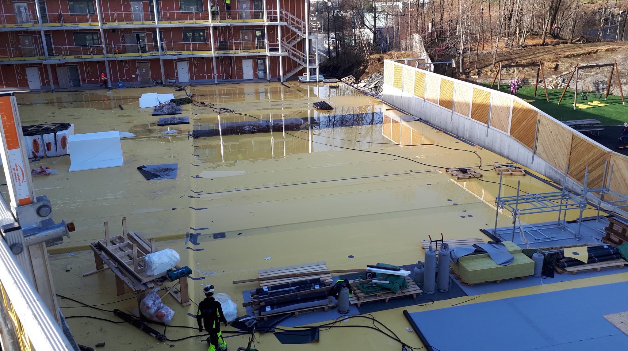 Budowa nowego budynku wielorodzinnego, widok z góry na żółtą izolację dachu, rusztowania, materiały budowlane i pracownika w kombinezonie ochronnym, w tle plac zabaw i drzewa.