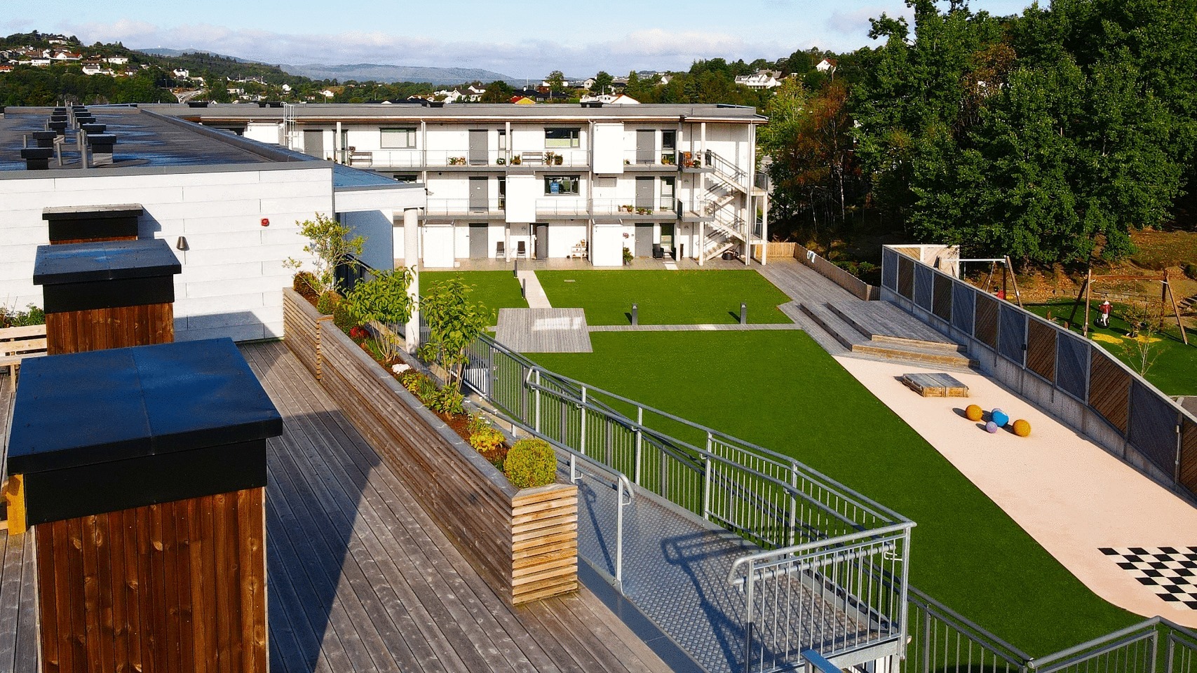 Widok z góry na nowoczesny kompleks mieszkalny z zielonym dziedzińcem, placem zabaw i drewnianym tarasem z roślinami w donicach.