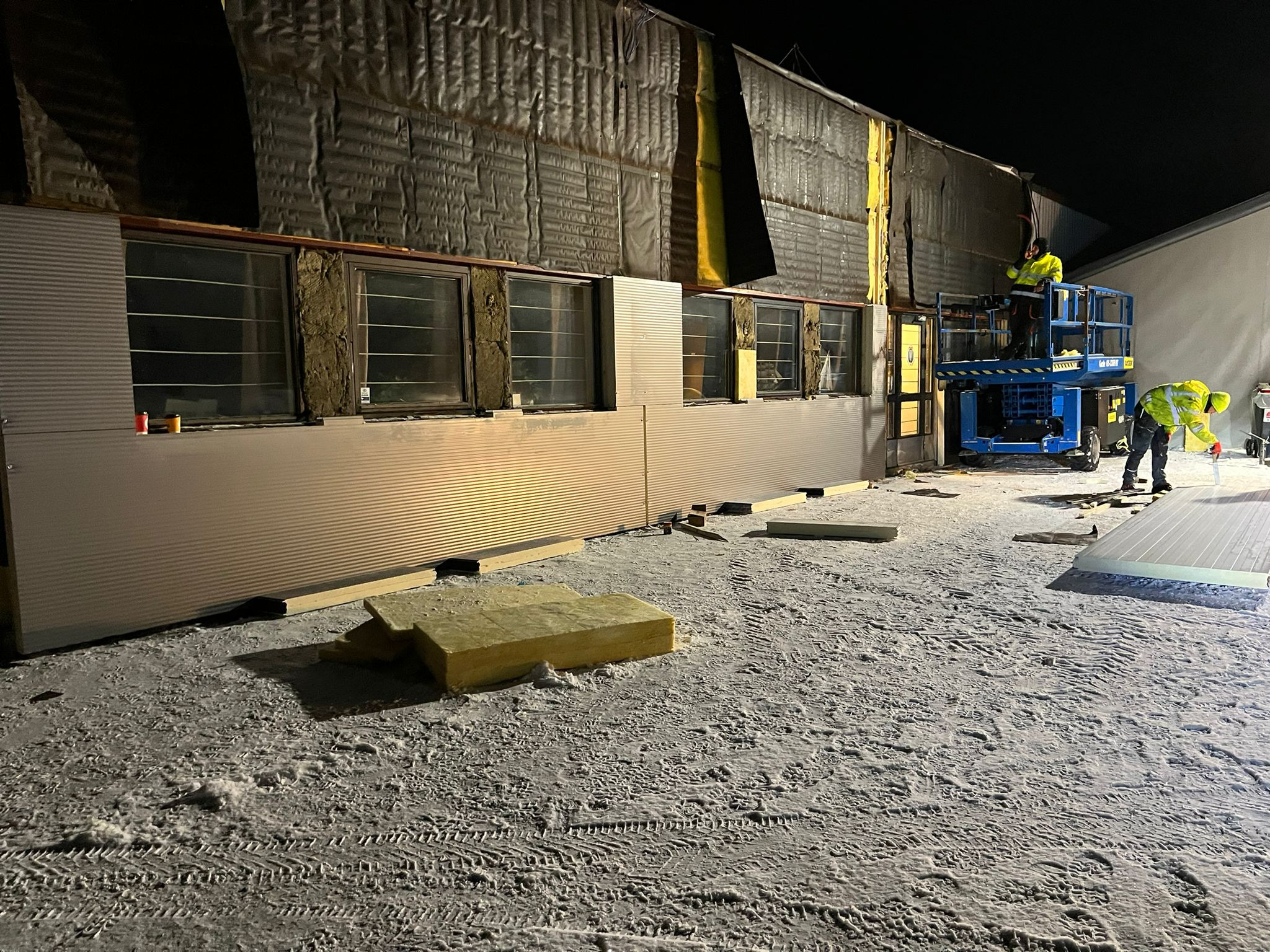 Montaż paneli elewacyjnych na budynku w zimowych warunkach, pracownicy na podnośniku koszowym i na ziemi, widoczne ocieplenie budynku i przygotowane panele.