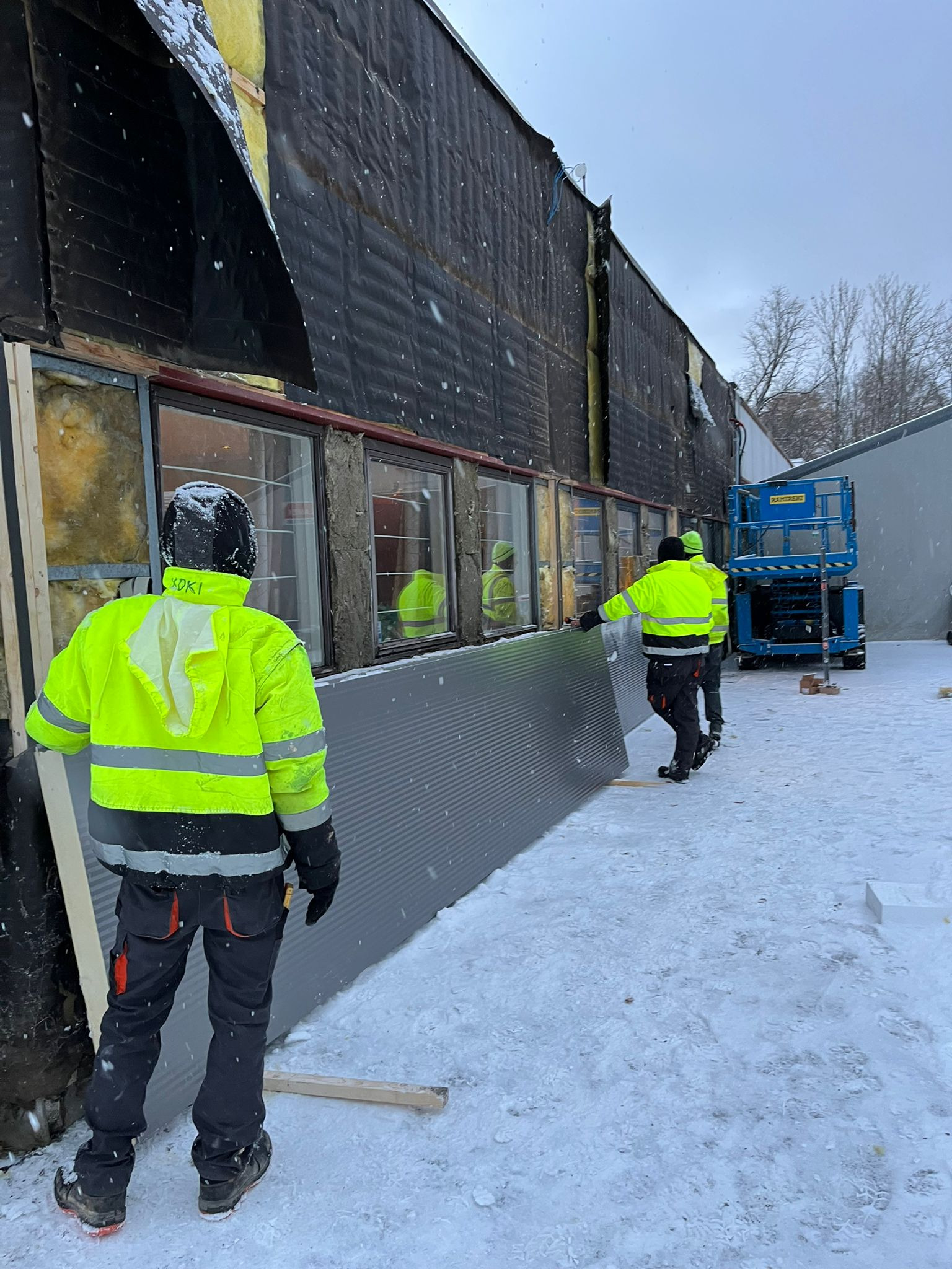 Montaż szarej blachy trapezowej na ścianie budynku z widoczną izolacją, pracownicy w odblaskowych kurtkach, zimowa sceneria i podnośnik koszowy w tle.