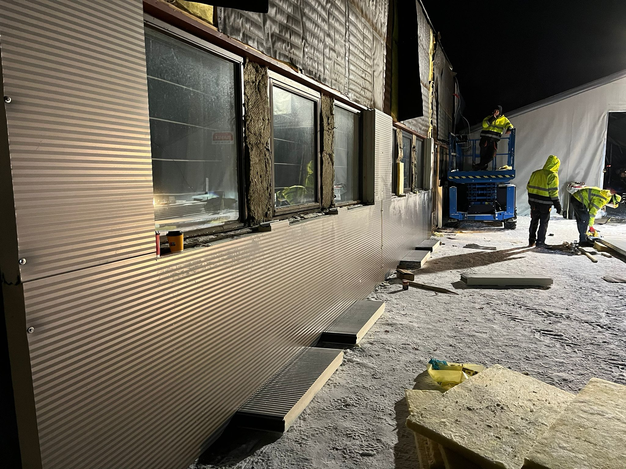 Montaż paneli ściennych na budynku, widoczni pracownicy w odblaskowych ubraniach na podnośniku oraz przy ścianie, nocna sceneria z ośnieżonym podłożem.