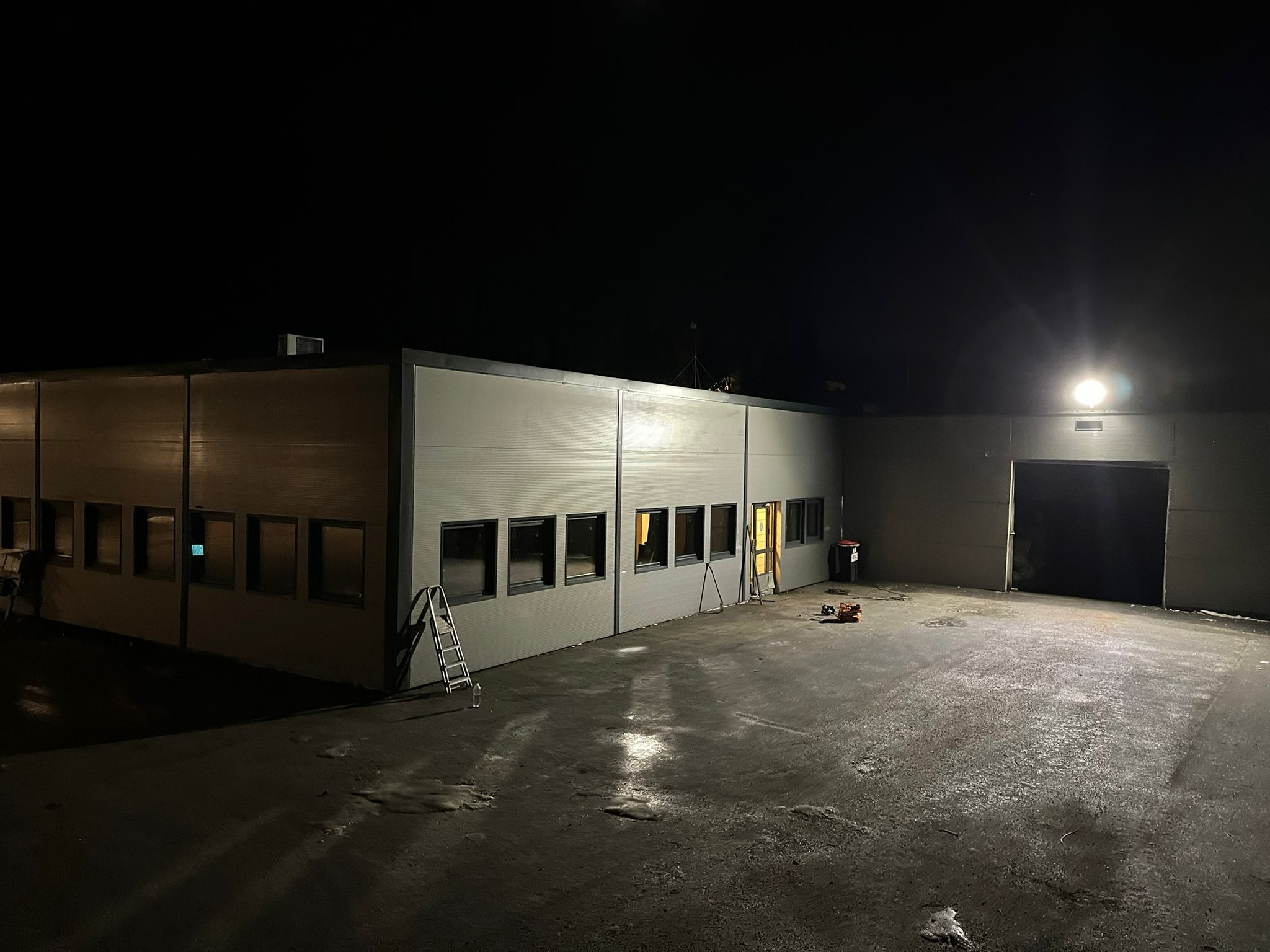 Nocne ujęcie elewacji budynku przemysłowego z widocznymi oknami, drabiną opartą o ścianę, śladami wilgoci na placu i narzędziami budowlanymi przy otwartych drzwiach.