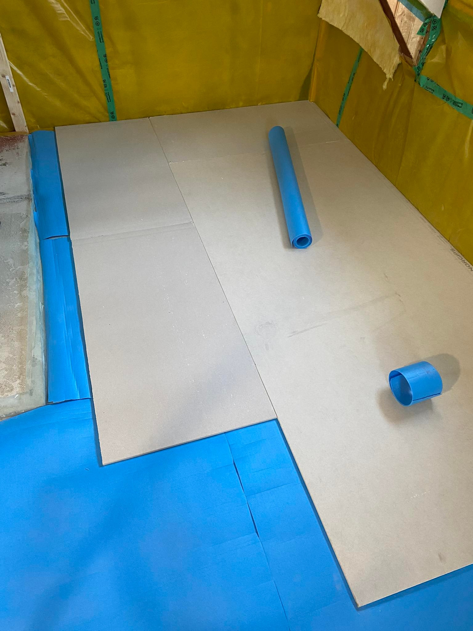 Przygotowanie podłoża pod płytę karton gips: rozłożona niebieska folia izolacyjna i płyty gipsowe na podłodze, widoczny zwinięty kawałek folii i odcięty kawałek rury z folii.