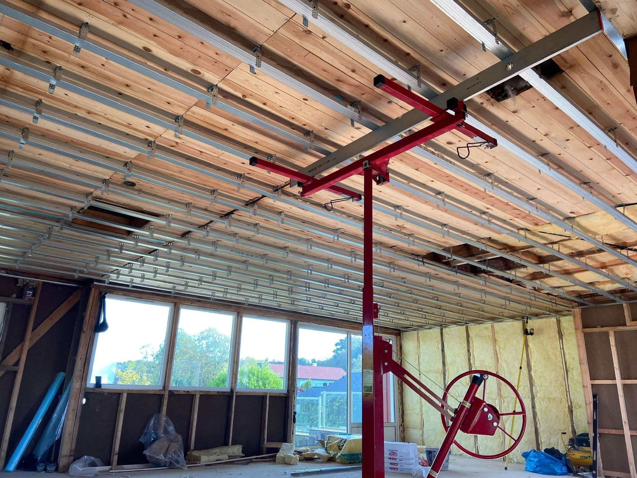 Montaż profili pod płytę karton gips na drewnianym suficie, widoczny podnośnik do płyt KG, ocieplenie ścian wełną mineralną, okna z widokiem na zewnątrz.