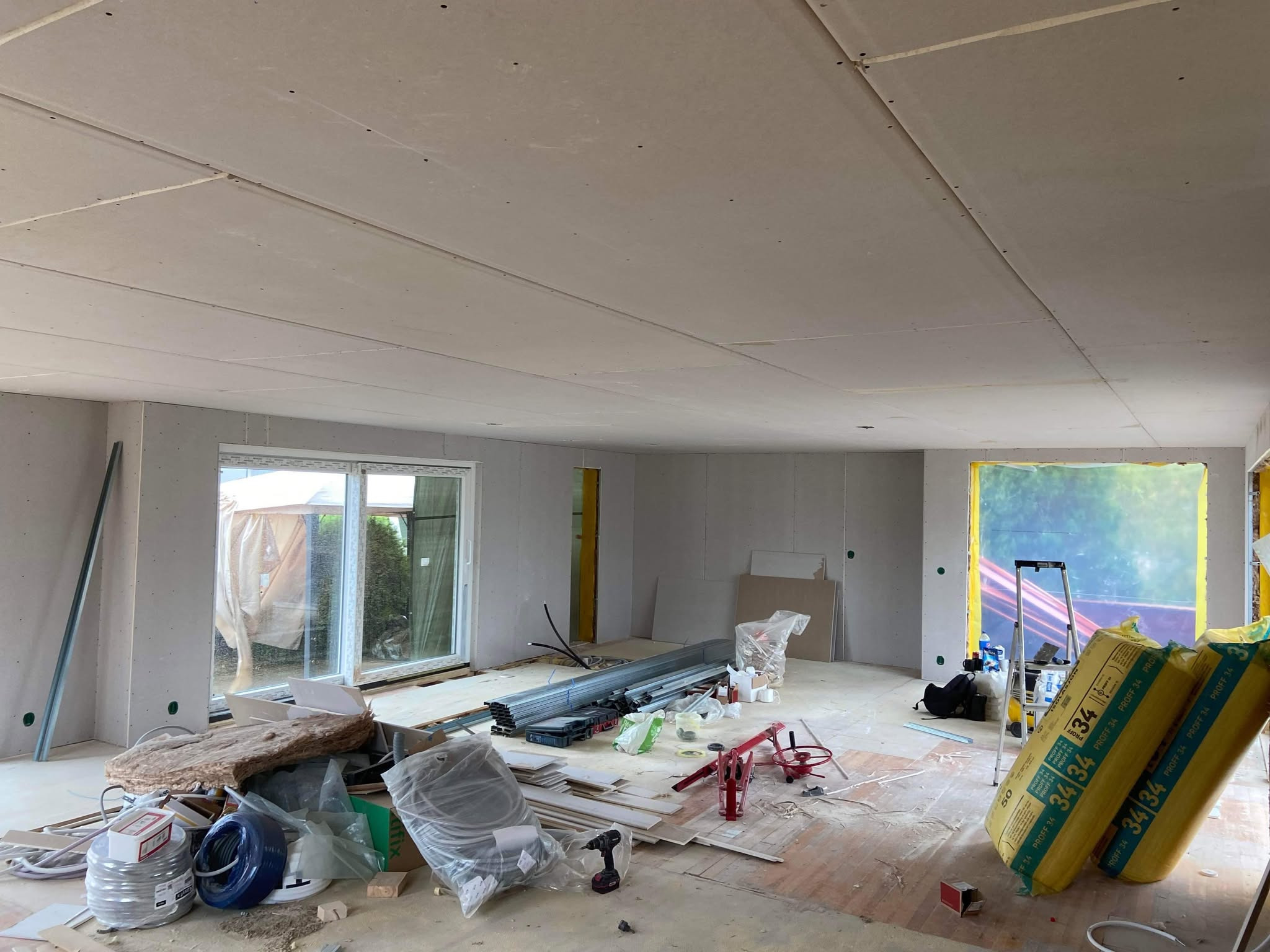 Wnętrze w trakcie montażu płyt kartonowo-gipsowych na ścianach i suficie, widoczne profile metalowe, narzędzia, materiały izolacyjne i otwory okienne.