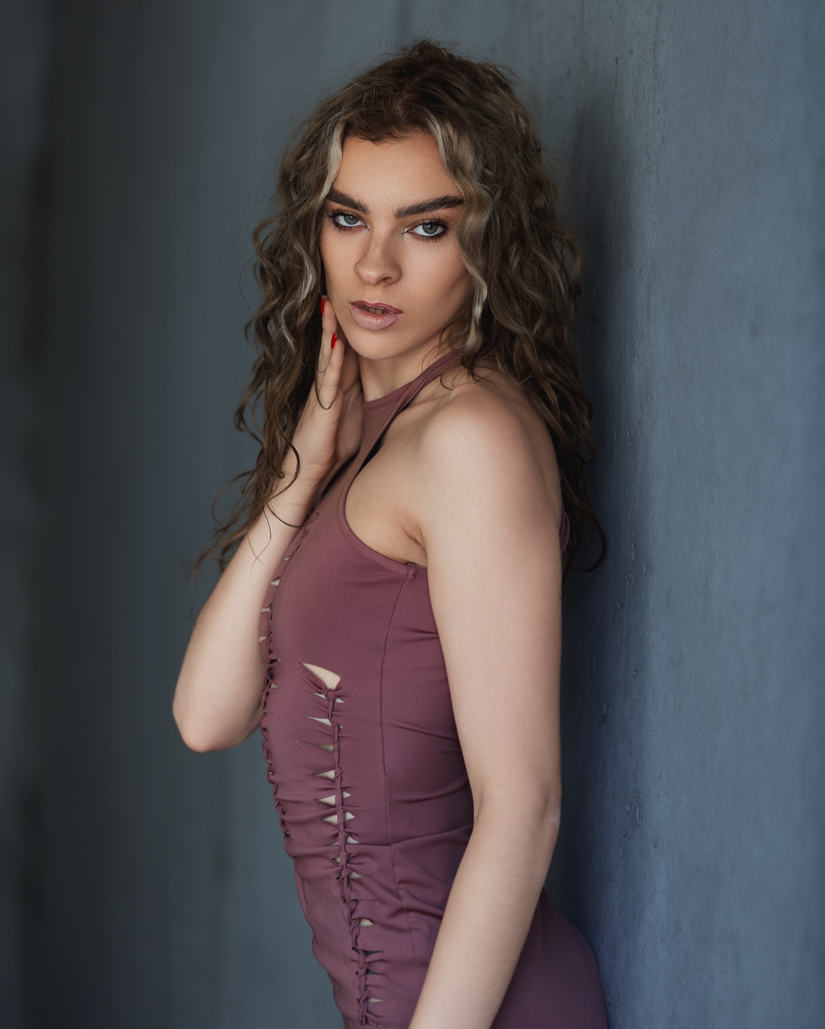 Portret kobiety z kręconymi włosami w dopasowanej sukience z wycięciami, opierającej się o szarą ścianę.