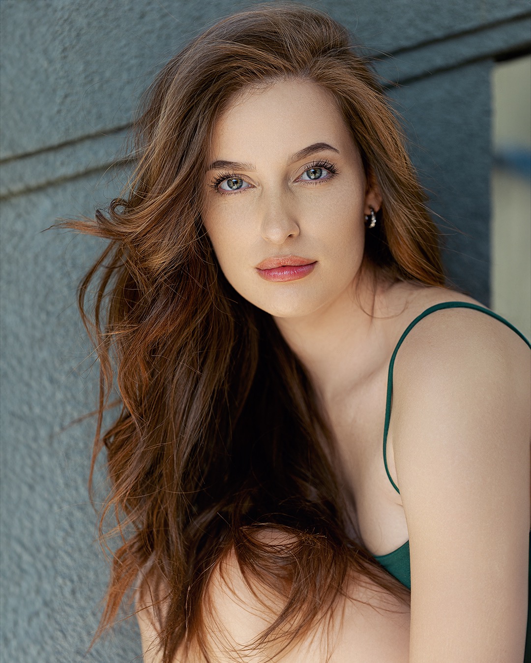 Portret kobiety z długimi, brązowymi włosami, w zielonej sukience na tle szarej ściany z wyraźną teksturą.