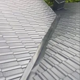 Błyszczący, grafitowy dach z dachówki ceramicznej, połączony z ciemną obróbką blacharską i kominem. Widok z góry, na tle zieleni drzew.