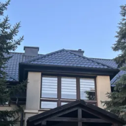 Dom jednorodzinny z ciemnym dachem krytym dachówką, widoczny z dołu, otoczony drzewami iglastymi i błękitnym niebem w tle. Widoczne okna z żaluzjami.