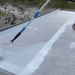 MARDAR Martyna Kujawa - Biały wałek malarski z długim uchwytem maluje szary dach. Część dachu jest już pomalowana na biało. Widoczne buty osoby malującej dach.