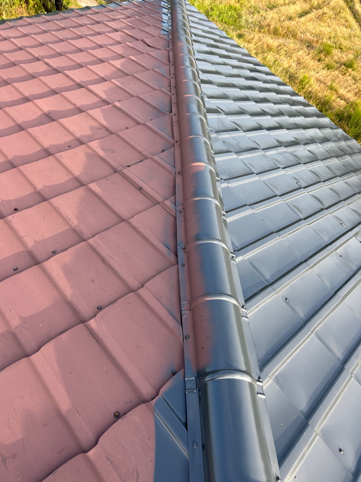 Dach z blachodachówki, połowa pomalowana na szaro, druga na czerwono. Widoczny łącznik kalenicowy. Trawa w tle.