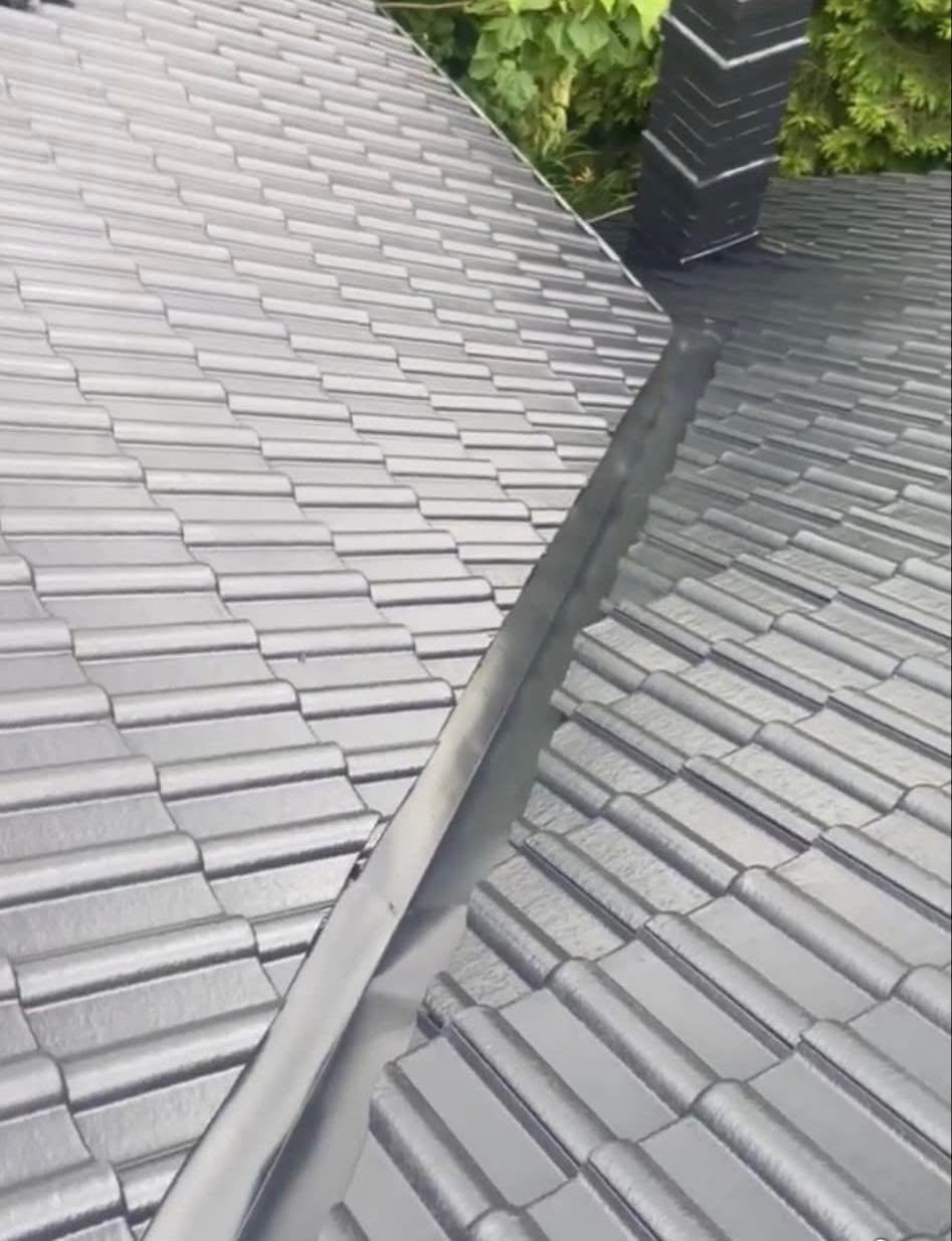 Błyszczący, grafitowy dach z dachówki ceramicznej, połączony z ciemną obróbką blacharską i kominem. Widok z góry, na tle zieleni drzew.