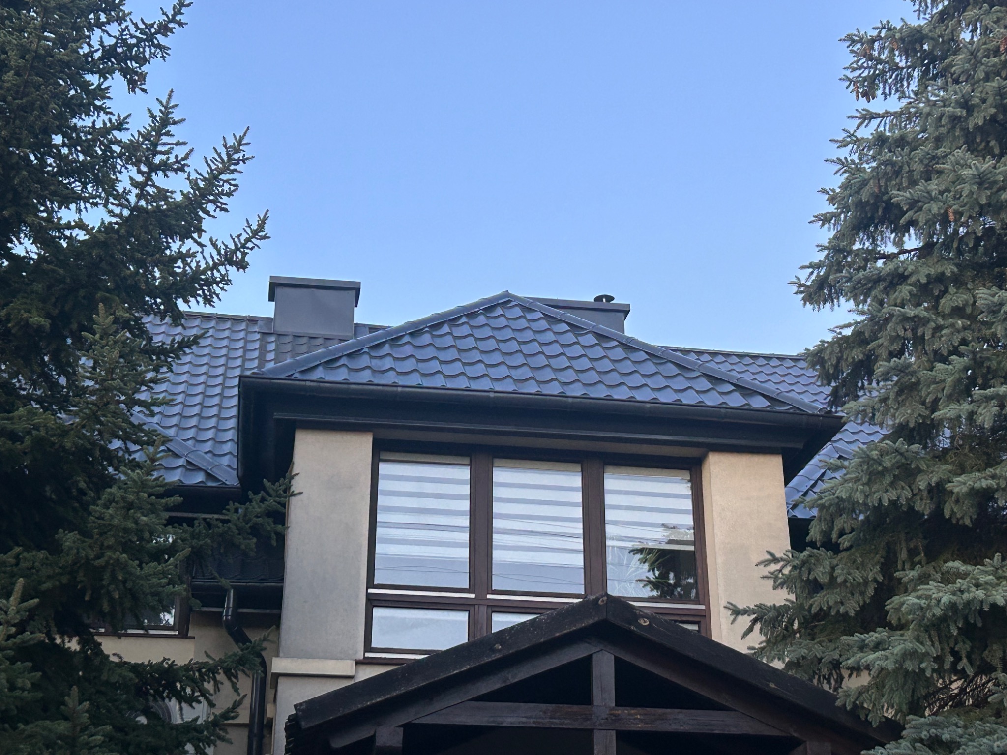 Dom jednorodzinny z ciemnym dachem krytym dachówką, widoczny z dołu, otoczony drzewami iglastymi i błękitnym niebem w tle. Widoczne okna z żaluzjami.