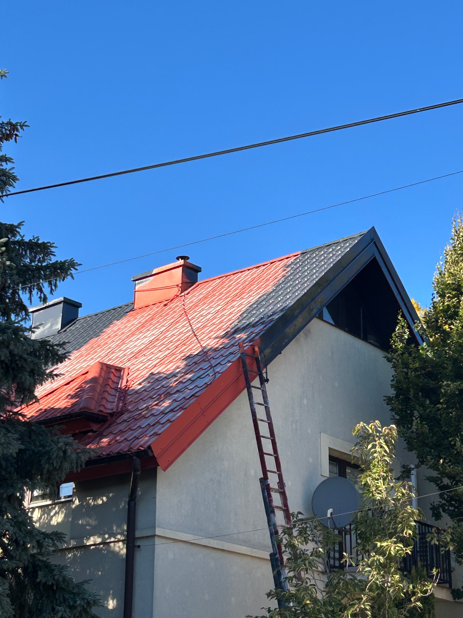Częściowo pomalowany dach z czerwonej blachodachówki, drabina oparta o ścianę domu, błękitne niebo w tle. Widoczne drzewa i antena satelitarna.