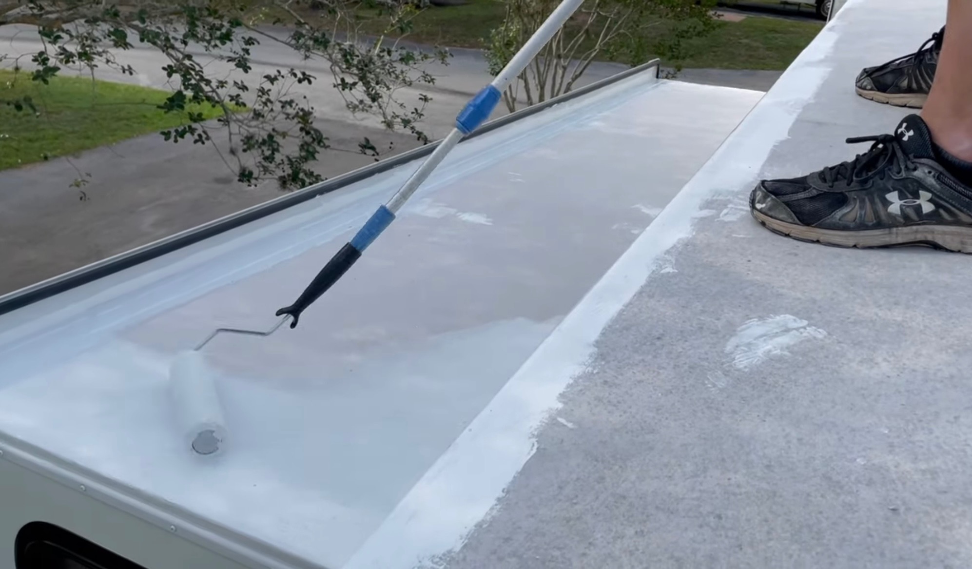 Biały wałek malarski z długim uchwytem maluje szary dach. Część dachu jest już pomalowana na biało. Widoczne buty osoby malującej dach.