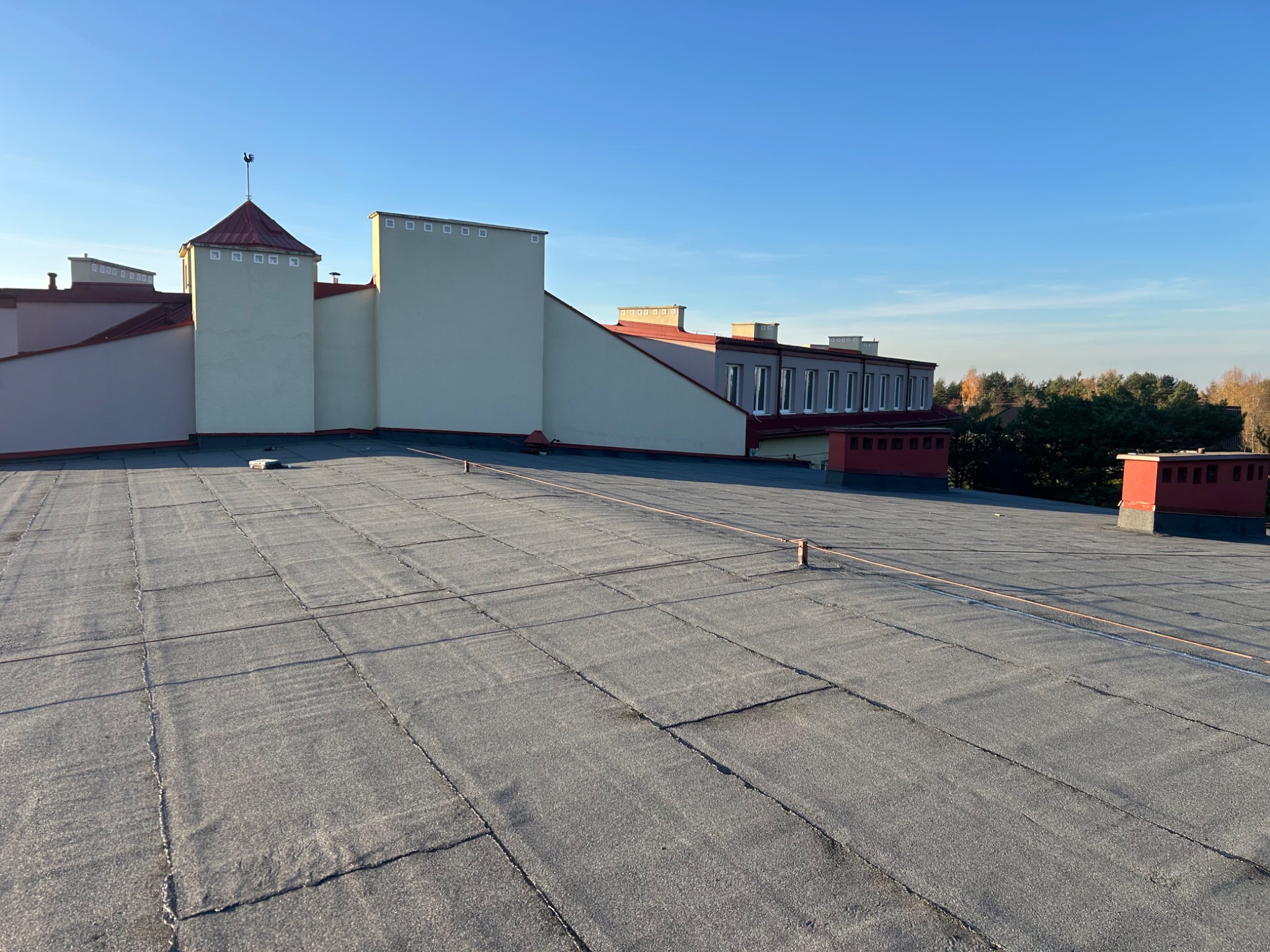 Szary, płaski dach budynku z widocznymi łączeniami papy, kominami i wieżyczką z wiatrowskazem na tle błękitnego nieba i drzew. Widok z góry, oświetlenie słoneczne.