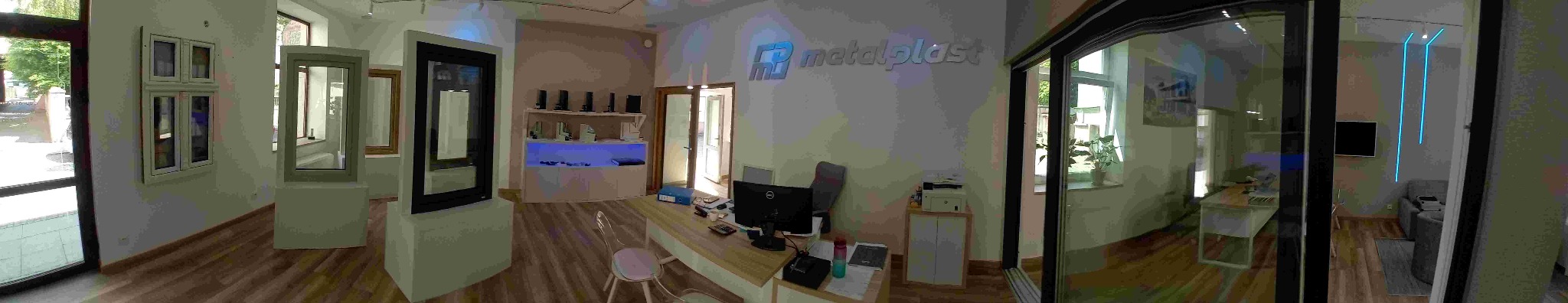 Ekspozycja okien i drzwi w salonie firmy Metalplast. Widoczne różne modele okien i drzwi, w tym przesuwne, na tle biura z logo firmy na ścianie.