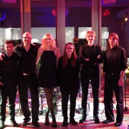 SINGERTON TOMASZ ŚPIEWAK - Zespół muzyczny w czarnych strojach pozuje na scenie z instrumentami, oświetlenie sceniczne w odcieniach czerwieni i fioletu, widoczny keyboard Korg.