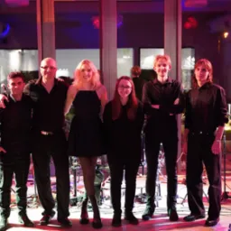 Zespół muzyczny w czarnych strojach pozuje na scenie z instrumentami, oświetlenie sceniczne w odcieniach czerwieni i fioletu, widoczny keyboard Korg.