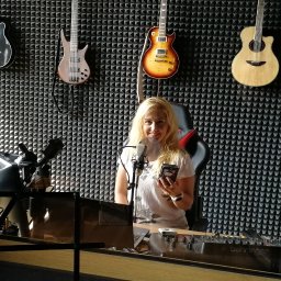 SINGERTON TOMASZ ŚPIEWAK - Kobieta z blond włosami uśmiecha się w studio nagraniowym, trzymając telefon, z gitarami wiszącymi na ścianie z panelami akustycznymi.