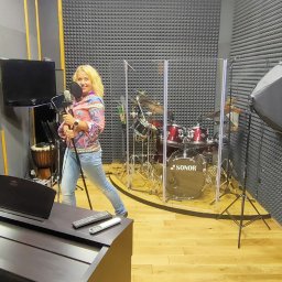 SINGERTON TOMASZ ŚPIEWAK - Kobieta z mikrofonem w ręku, w profesjonalnym studio nagrań z pianinem, perkusją za plecami i panelami akustycznymi na ścianach.