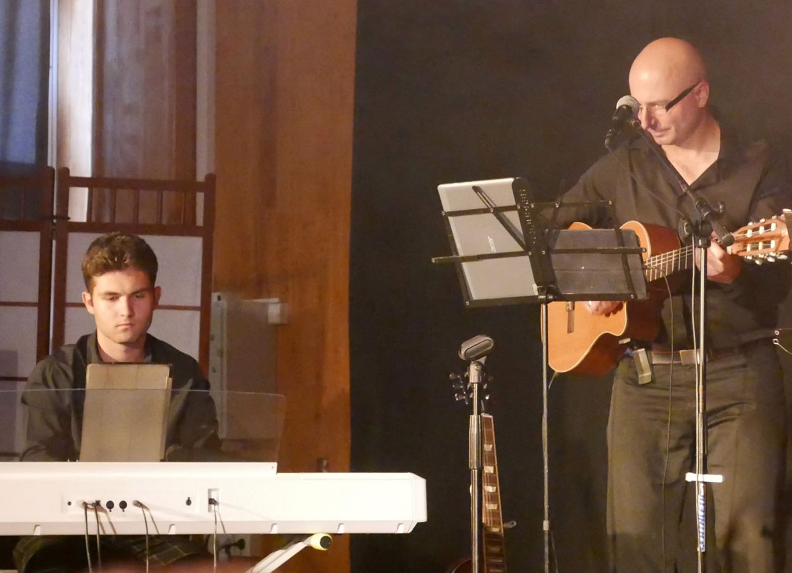 Mężczyzna z gitarą śpiewa do mikrofonu, obok drugi muzyk gra na keyboardzie, dodatkowo widoczna gitara basowa na statywie.