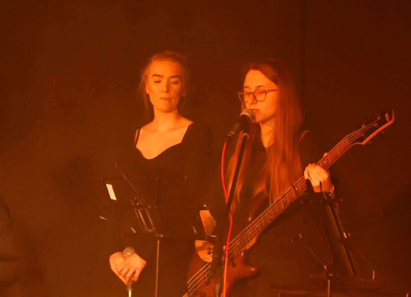 Dwie kobiety na scenie podczas występu: jedna śpiewa do mikrofonu grając na gitarze basowej, druga stoi obok z mikrofonem i nutami, oświetlone ciepłym, pomarańczowym światłem.