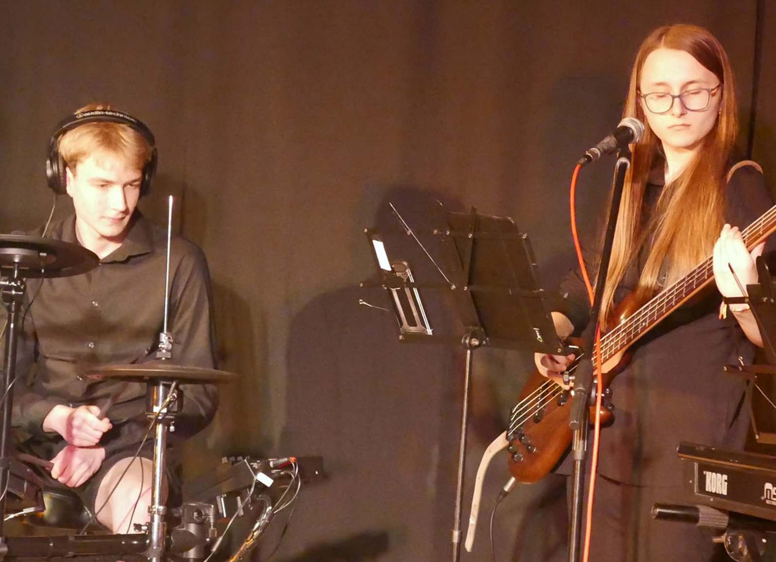 Młody perkusista w słuchawkach gra na perkusji elektronicznej, obok basistka z długimi włosami i okularach gra na basie na tle ciemnej ściany.