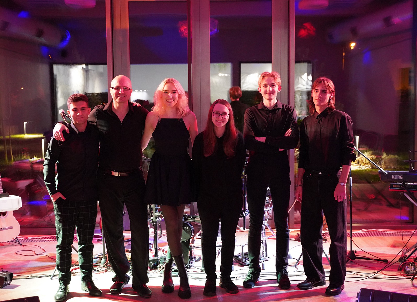 Zespół muzyczny w czarnych strojach pozuje na scenie z instrumentami, oświetlenie sceniczne w odcieniach czerwieni i fioletu, widoczny keyboard Korg.