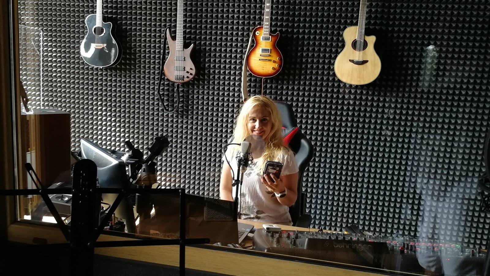 Kobieta z blond włosami uśmiecha się w studio nagraniowym, trzymając telefon, z gitarami wiszącymi na ścianie z panelami akustycznymi.